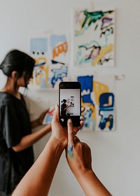 Friend taking a photo with phone camera of her artist friend by a mood board on a white wall