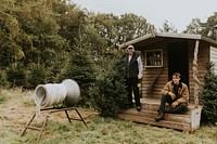 Two men by a Christmas wrapping tube machine and a log cabin 