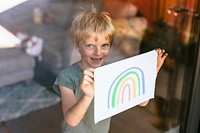 Blond boy showing rainbow on paper through glass window, the new normal