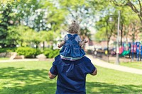 Little girl rides on father's shoulders on the grass.