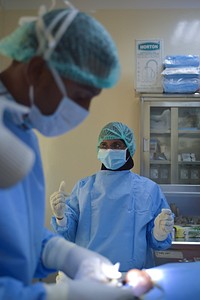 A nurse stands in the operating room of the Sheikh Zayed Bin Sultan Al'Nahya Hospital in Mogadishu, Somalia, while a doctor performs minor surgery in order to remove a lymphatic cyst. AMISOM Photo / Tobin Jones. Original public domain image from <a href="https://www.flickr.com/photos/au_unistphotostream/20586766132/" target="_blank" rel="noopener noreferrer nofollow">Flickr</a>
