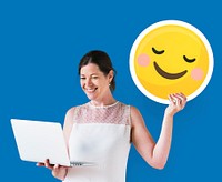 Woman holding a blushing emoticon and using a laptop