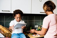 Black kid using tablet while mother cooking in the kitchen