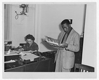 [Gordon Parks, Farm Security Administration/Office of War Information photographer, standing in office with Helen Wool seated at desk]. Sourced from the Library of Congress.