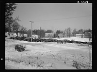 Bantam, Connecticut. In the background is the Warren McArthur plant. The cars in the foreground belong to workers in the plant, at least half of whom commute from nearby towns and cities. Between the cars and the bridge runs a track along which, once daily, a train pulls up with feed and grain for Bantam's daily farmers. Sourced from the Library of Congress.