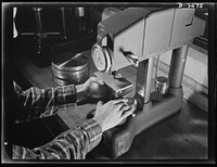 Conversion. Copper and brass processing. This sample of brass strip is being tested on a Rockwell Hardness Testing Machine. This test is based upon the depth a steel ball penetrates the metal. A minor load is applied first to hold the ball in position, and then a major load is applied. The dial on the machine measures the depth of the penetration. This test is made to check the physical properties of the metal. Chase Copper and Brass Company, Euclid, Ohio. Sourced from the Library of Congress.