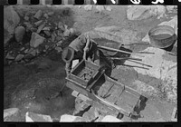 Eugene Davis, gold prospector, operating "rocker". Rocks and sticks remain in the screened box at the top when water is sluiced over them and the contraption is rocked by the handle. Loose dirt and any mineral content washes below, the dirt gradually washing through and the heavy gold and minerals settling to the bottom and behind the ridges. Pinos Altos, New Mexico by Russell Lee