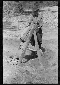 Prospector operating dry washer called a papago. This dry washer is the old Indian method of washing for gold. The rocks and dirt are placed in the higher compartment. Below and to the back is a bellows made of canvas. This is pumped to blow through the screening and thus blow away loose dirt while the heavy gold remains. Pinos Altos, New Mexico. by Russell Lee