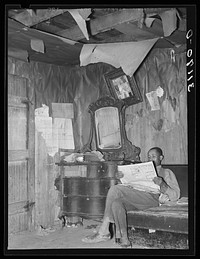 Sharecropper reading newspaper in corner of living room. Note the bureau and ceiling. Near Southeast Missouri Farms by Russell Lee