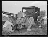 Migrant steeple-jack dropping the pan of his automobile before replacing burned-out bearing. His father and wife are also shown. When he secures a job, the entire family, consisting of his father and mother, wife and sister, all help with the painting. Camped near Prague, Oklahoma by Russell Lee