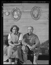  preacher and his wife sitting under photos taken of them twenty years ago. They live in an old converted schoolhouse with two grandchildren. The rest of their children have moved out of the county. Heard County, Georgia. Sourced from the Library of Congress.