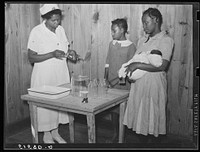 Nurse Shamburg demonstrates care of baby bottles to Mrs. Paralee Coleman and Marie. Mothers have to work in field and children are left at home to care for babies. Health clinic, Gee's Bend, Alabama. Sourced from the Library of Congress.