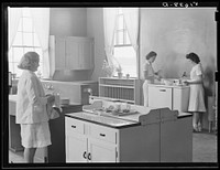 Margaret Segars at sink with dinner tray showing well-balanced meal at right. In background are Dorothy Smith and Elizabeth Atkinson cooking on electric stove in home economics class room in school building. Ashwood Plantations, South Carolina. Sourced from the Library of Congress.