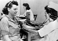The 1950s historical photograph of a nurse taking her blood pressure of a woman ordnance worker in the hospital.
