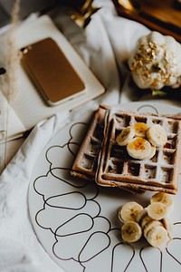 Plate of breakfast waffle with banana topping on a white bed next to a diary and a phone