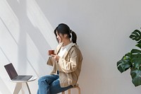Happy Asian woman sitting on a wooden stool drinking coffee while using her laptop 