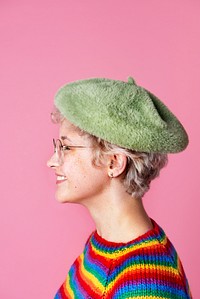 Side view of a cute girl wearing a rainbow sweater and a green beret in a pink background