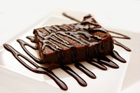 A chocolate brownie on a white plate decorated with chocolate drizzle. Original public domain image from <a href="https://commons.wikimedia.org/wiki/File:Brownie-dessert-cake-sweet-45202.jpg" target="_blank" rel="noopener noreferrer nofollow">Wikimedia Commons</a>