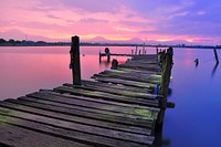 An old dock that is falling apart over the water that is reflecting the pink and purple sky. Original public domain image from Wikimedia Commons