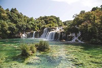 People swimming in a lake under a wide waterfall on a sunny summer's day. Original public domain image from Wikimedia Commons