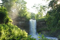 A small waterfall pouring down from a rocky ledge in a sunny forest. Original public domain image from Wikimedia Commons