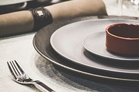 A red soup bowl on black and gray ceramic plates beside a shiny silver fork and a rolled up napkin. Original public domain image from Wikimedia Commons