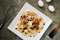 Banana pancakes with raisins and syrup on a white plate, eggs, and cooking utensils on a countertop in Makati. Original public domain image from Wikimedia Commons