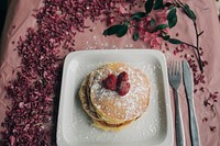 Delicious looking pancakes covered in berries and powdered sugar on a plate that is sitting on a table with flowers on it. Original public domain image from Wikimedia Commons