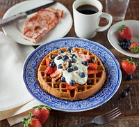 Round waffles with whipped cream, strawberries and blueberries next to a cup of coffee. Original public domain image from <a href="https://commons.wikimedia.org/wiki/File:Fruit_waffles_and_coffee_(Unsplash).jpg" target="_blank" rel="noopener noreferrer nofollow">Wikimedia Commons</a>
