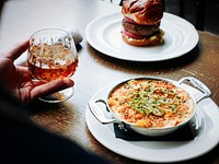 Hand holding a cocktail next to a cheese and jalapeno dip and a burger. Original public domain image from Wikimedia Commons