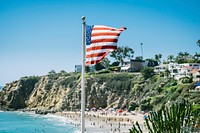 The American flag flutters in the wind over a crowded beach by a cliff. Original public domain image from <a href="https://commons.wikimedia.org/wiki/File:Flag_over_a_beach_(Unsplash).jpg" target="_blank" rel="noopener noreferrer nofollow">Wikimedia Commons</a>