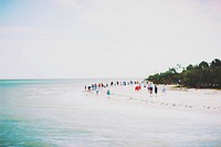 People dressed in an array of bright colors stand along the blue sea shore.. Original public domain image from Wikimedia Commons