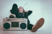 A blurry shot of a woman in sneakers reclining with her foot on a vintage sound system. Original public domain image from <a href="https://commons.wikimedia.org/wiki/File:Boombox_(Unsplash).jpg" target="_blank" rel="noopener noreferrer nofollow">Wikimedia Commons</a>