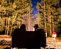 Two people seated in a sofa near a campfire in a forest at night. Original public domain image from <a href="https://commons.wikimedia.org/wiki/File:A_Couple_in_Glow_(Unsplash).jpg" target="_blank" rel="noopener noreferrer nofollow">Wikimedia Commons</a>