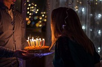 A man bringing a birthday cake with candles to a woman in a room filled with balloons. Original public domain image from <a href="https://commons.wikimedia.org/wiki/File:Happy_Birthday!_(Unsplash).jpg" target="_blank" rel="noopener noreferrer nofollow">Wikimedia Commons</a>