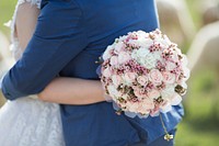 A bride embraces her groom while holding a beautiful floral bouquet behind his back. Original public domain image from <a href="https://commons.wikimedia.org/wiki/File:Floral_embrace_(Unsplash).jpg" target="_blank" rel="noopener noreferrer nofollow">Wikimedia Commons</a>