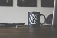 A black mug sitting on an office desk in Missoula with white writing which says "Hustle". Original public domain image from Wikimedia Commons