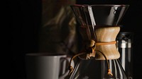 Chemex pour-over glass coffee maker next to ground coffee and a mug against dark background. Original public domain image from Wikimedia Commons