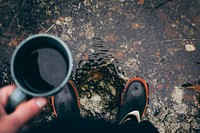 A person wearing rain boots stands in a puddle looking down and holding coffee. Original public domain image from Wikimedia Commons