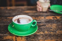 A latte in a green cup on a table with a person drinking their coffee on the other side. Original public domain image from Wikimedia Commons