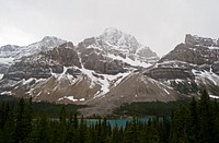On a cloudy winter day in Canada, snow covers a rocky mountain range flanked with pine trees surrounding a lake. Original public domain image from Wikimedia Commons
