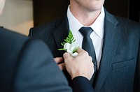 A groomsman in a suit pins a boutonniere on the groom, who wears a black tie. Original public domain image from Wikimedia Commons