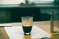 A glass cup of coffee placed on a paper on a table. Original public domain image from Wikimedia Commons