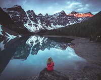 A woman in a red jacket sitting on a rock at the shore of a mountain lake. Original public domain image from Wikimedia Commons