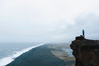 Man standing on a cliff overlooking a black sand shoreline in Vik. Original public domain image from Wikimedia Commons