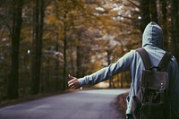 A hooded hitchhiker with a backpack extending his thumb on the side of an empty road. Original public domain image from Wikimedia Commons