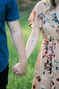 A man and a woman standing outdoors hold hands. She has a tattoo on her arm. Original public domain image from <a href="https://commons.wikimedia.org/wiki/File:Holding_hands_in_a_meadow_(Unsplash).jpg" target="_blank">Wikimedia Commons</a>