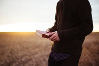 Man in sweater holding bible with two hands in a field during sunset. Original public domain image from Wikimedia Commons