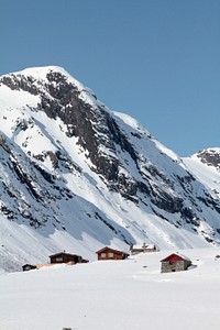 Houses at the top of a hill with snow capped mountains in the background and blue skies. Original public domain image from <a href="https://commons.wikimedia.org/wiki/File:Strynefjellet%27s_snowy_cabins_(Unsplash).jpg" target="_blank" rel="noopener noreferrer nofollow">Wikimedia Commons</a>