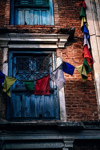 A brick building with blue window shutters and colored flags around it in Swayambhu Stupa. Original public domain image from Wikimedia Commons
